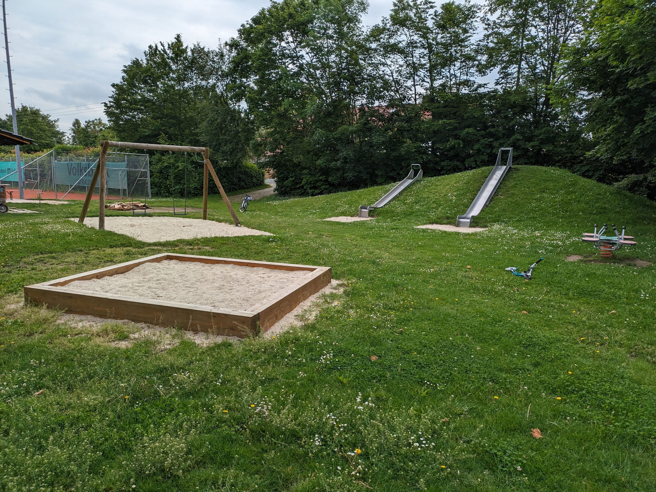 Spielplatz mit zwei Schaukeln, zwei Rutschen, einer Wippe und Sandkasten. Im Hintergrund ist ein Tennisplatz zu sehen